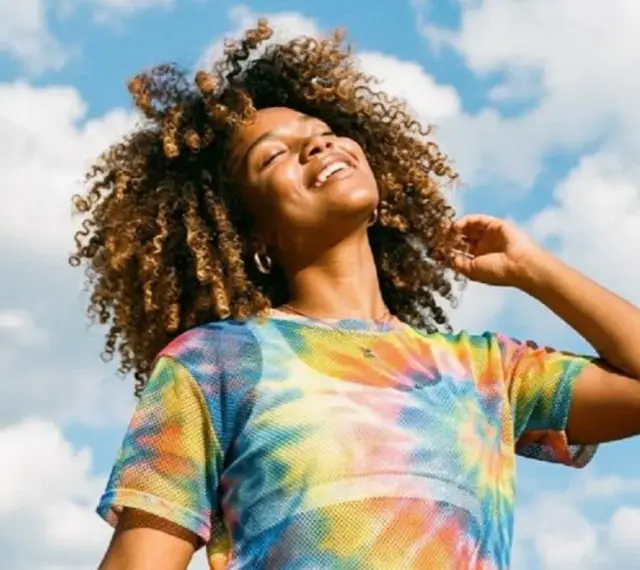 Une femme souriante aux cheveux bouclés, sous un ciel bleu nuageux. Elle porte un t-shirt tie-dye coloré. Une belle image pour apprendre comment rendre une photo flou.