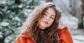 Jeune femme aux cheveux bouclés, portant une veste orange. La retouche photo netteté en ligne sublime son regard intense.