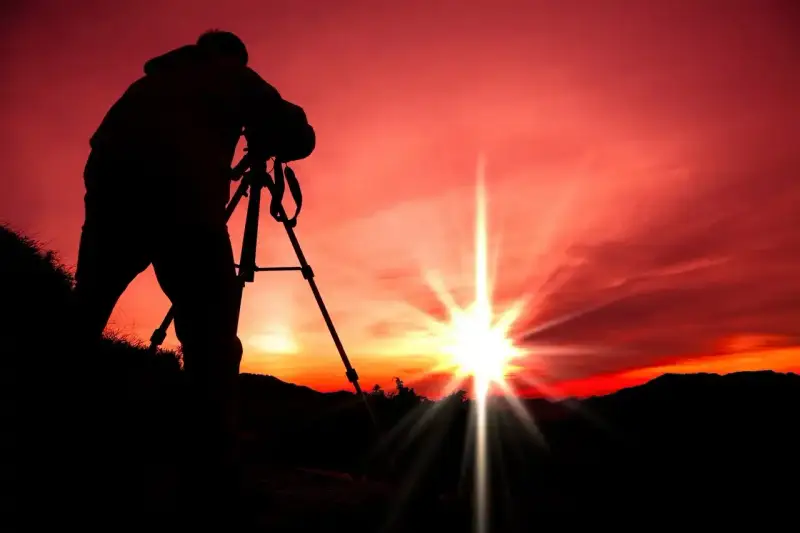 Silhouette d'un photographe sur trépied, capturant le soleil éclatant lors d'un coucher de soleil rougeoyant.
