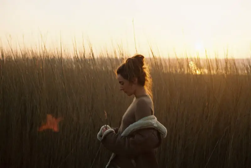 Une jeune femme, le regard baissé, se tient dans les hautes herbes au crépuscule. La lumière dorée du soleil couchant illumine ses cheveux et le paysage.